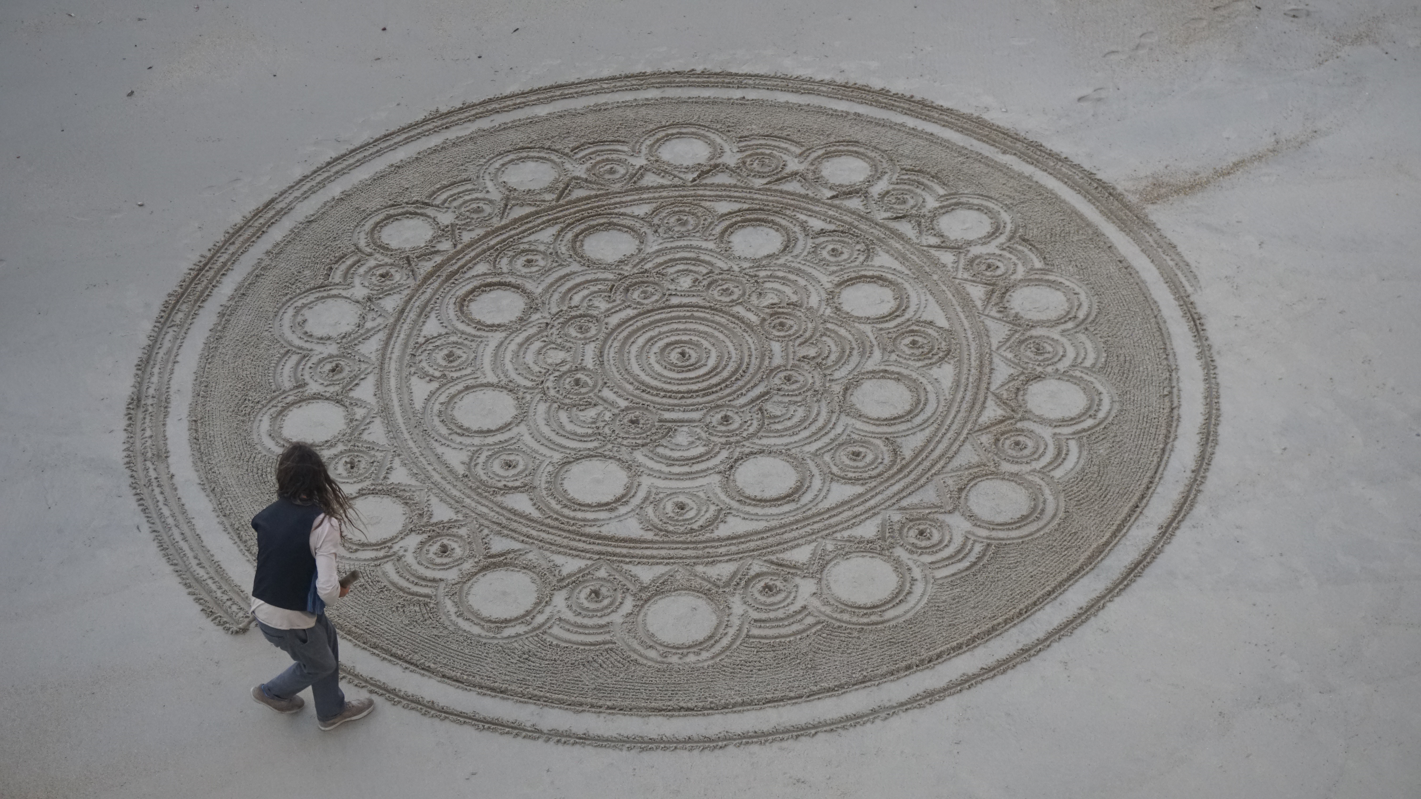A person beside a large circular pattern drawn in beach sand.