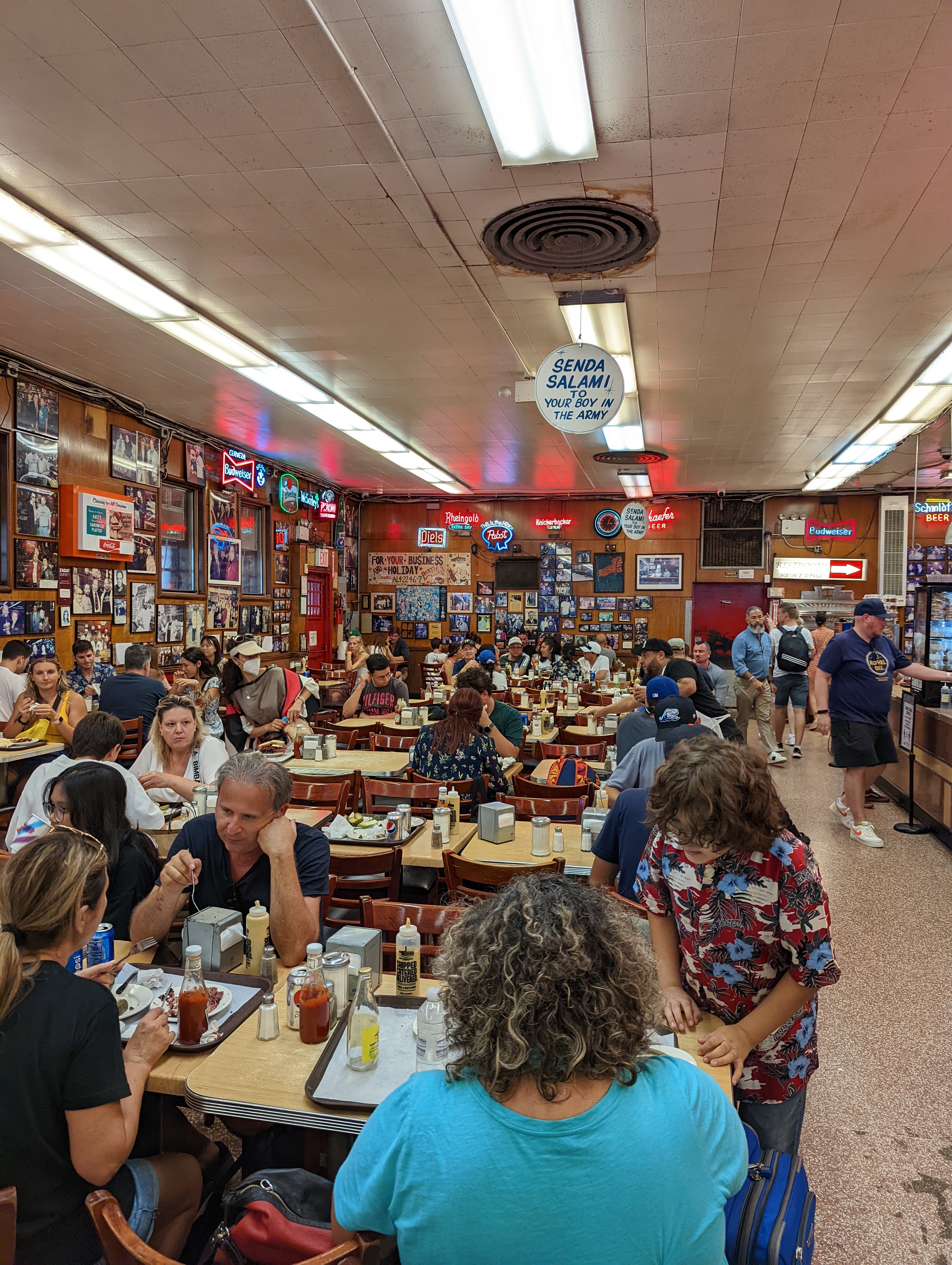 Diners sit at closely packed tables inside a deli, with framed photographs covering the walls.