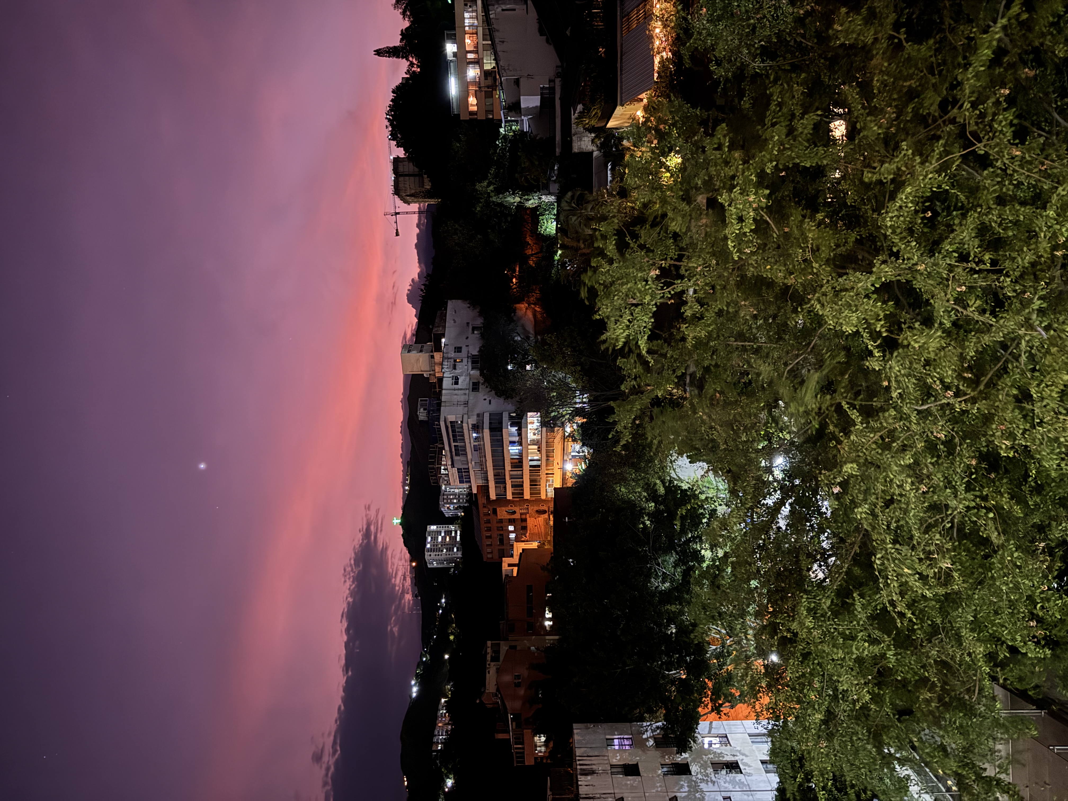 A violet and pink sky above trees, city buildings and dark hills.